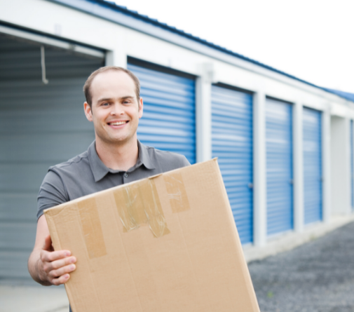A smiling man holding a cardboard moving box in front of a row of self storage units