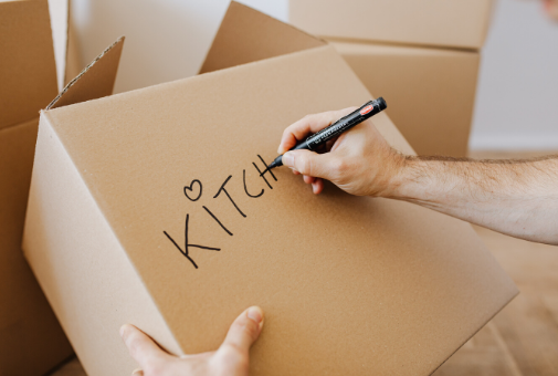 A hairy arm writing "Kitchen" on a moving box